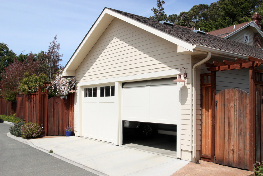 A beige garage with a partially open door, surrounded by a wooden fence and trees, next to a paved driveway.